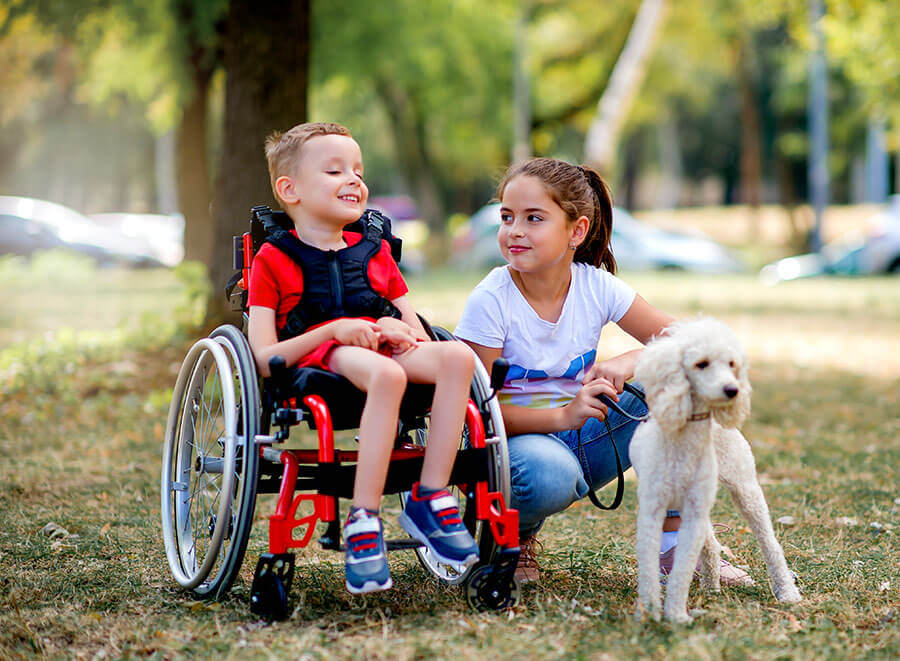 A boy and a girl in the park. The boy is sitting in a wheelchair and smiling. The girl is holding a dog on a lead.